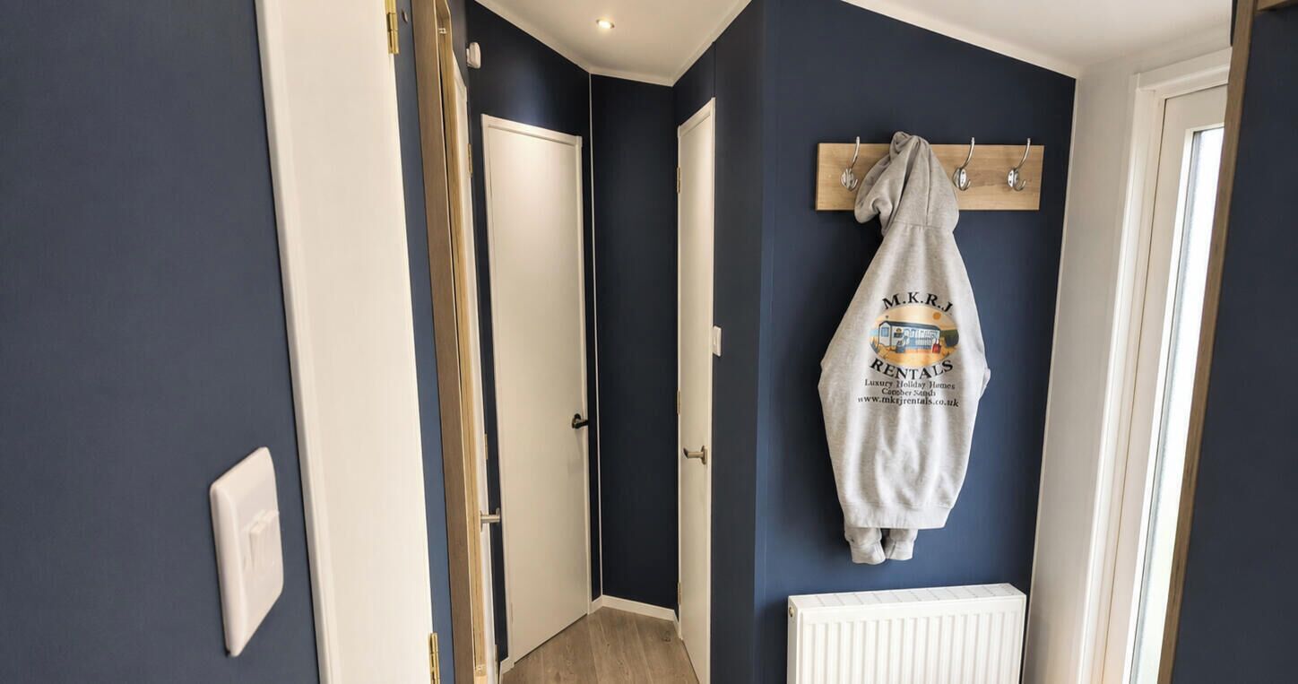 Un couloir avec un mur bleu foncé, une porte blanche, un banc en bois sur lequel sont posées des bottes jaunes, et un sweat-shirt gris accroché à un crochet.