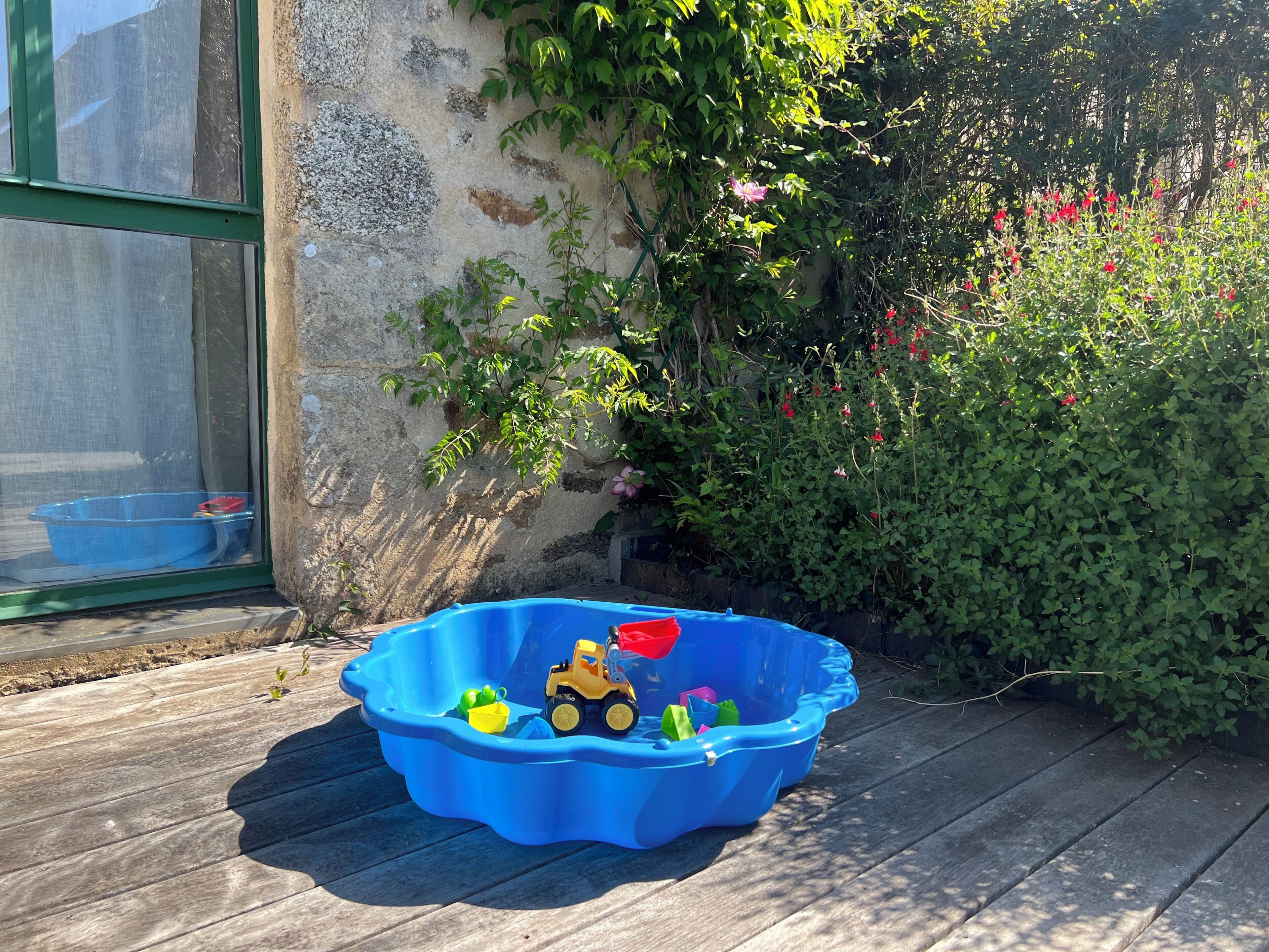 A blue plastic kiddie pool filled with toys and a small yellow toy vehicle on a wooden deck.