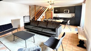 Uma sala de estar ampla com um sofá, duas poltronas, uma mesa de centro e uma cozinha ao fundo.