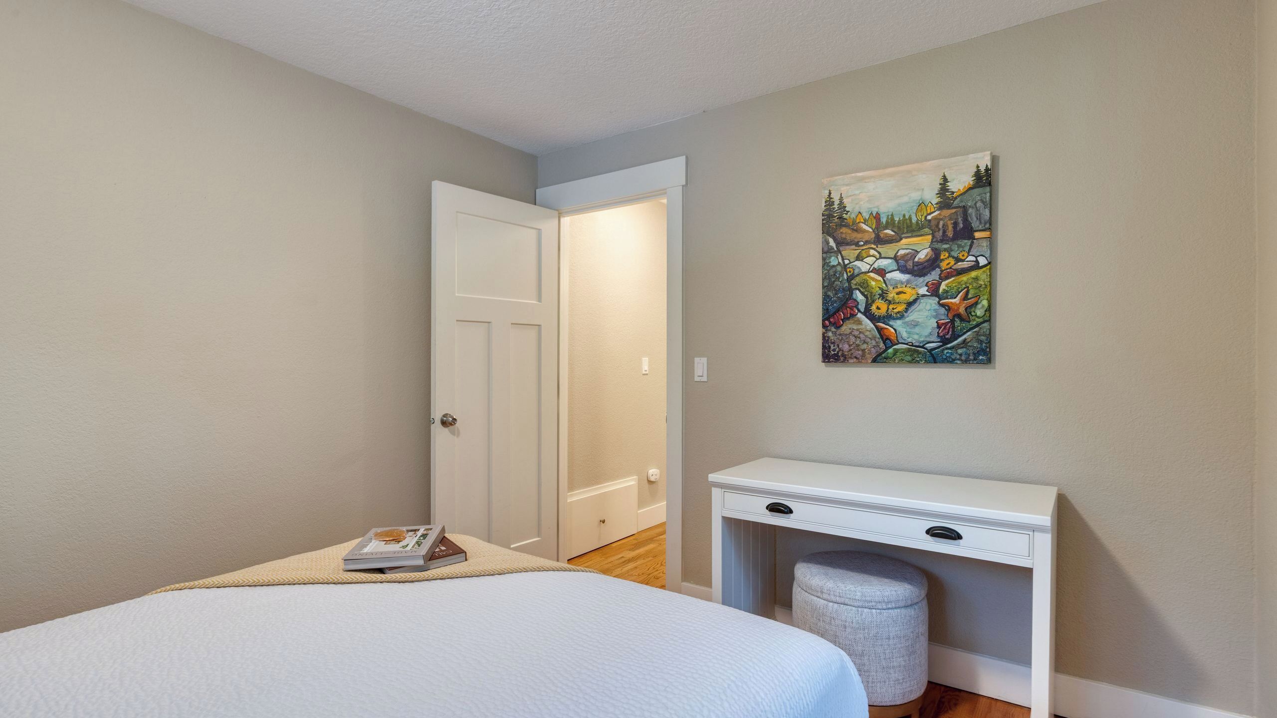 Um quarto com uma cama, uma escrivaninha, uma cadeira, um quadro e uma porta.