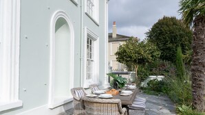 Eine Terrasse mit Korbmöbeln, einem Steintisch gedeckt für ein Essen und ein weißes Gebäude mit Rundbogenfenstern.