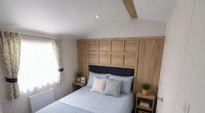 Een slaapkamer met een houten hoofdbord, een bed met veel kussens, een nachtkastje met een plant en een raam met gordijnen.