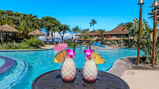 Ein Poolbereich mit Tiki-Getränken, Palmen und blauem Himmel.