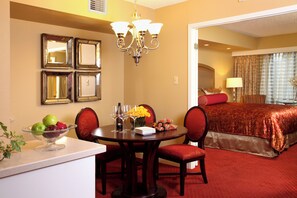 Ein Hotelzimmer mit Essbereich, der einen runden Tisch, Stühle und eine Obstschale zeigt. Über dem Tisch hängt eine Kristallleuchter, im Hintergrund ist ein Bett mit einer roten Tagesdecke zu sehen.