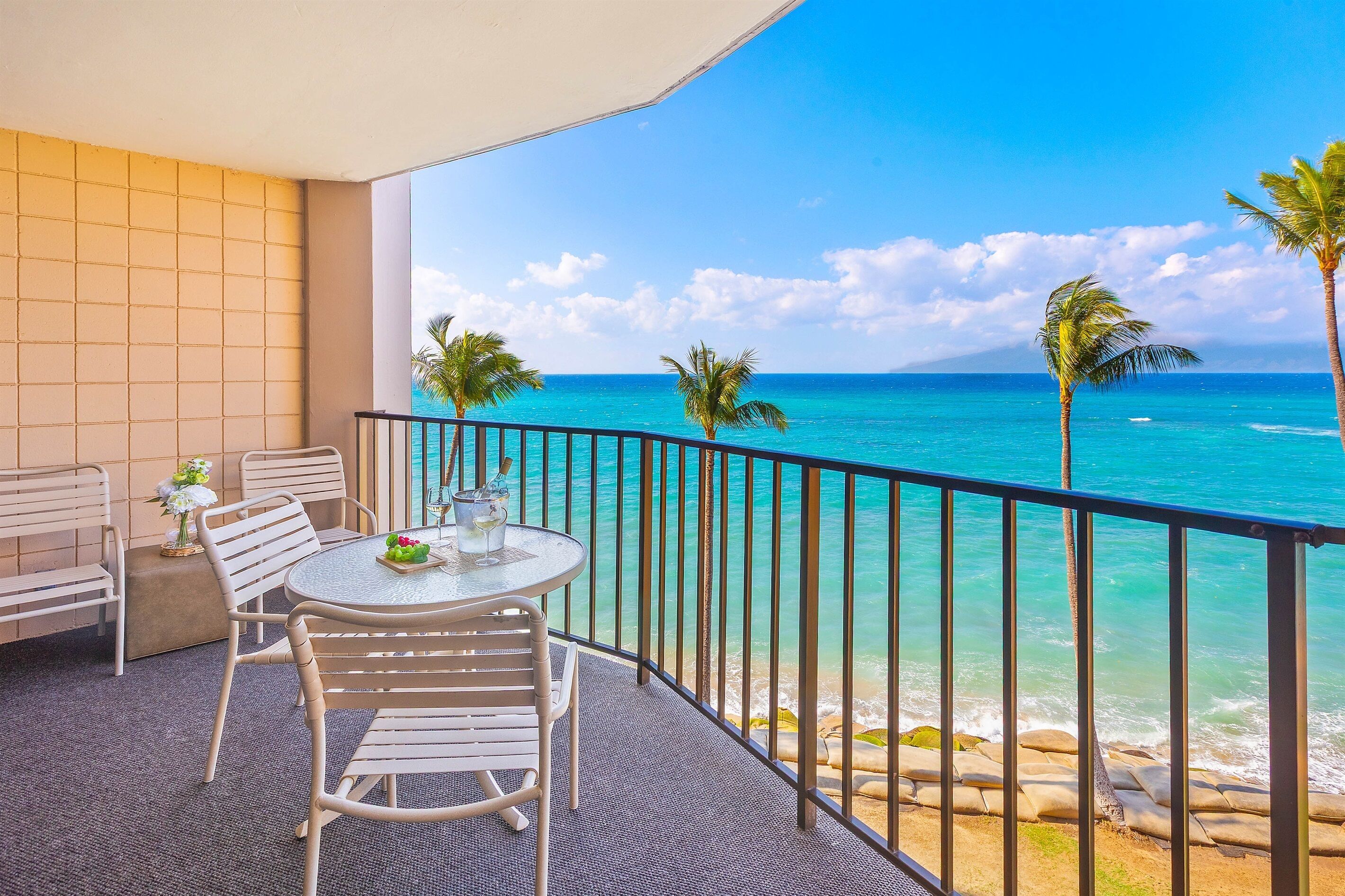 A balcony with a table and chairs, overlooking a beach with clear blue water and palm trees.