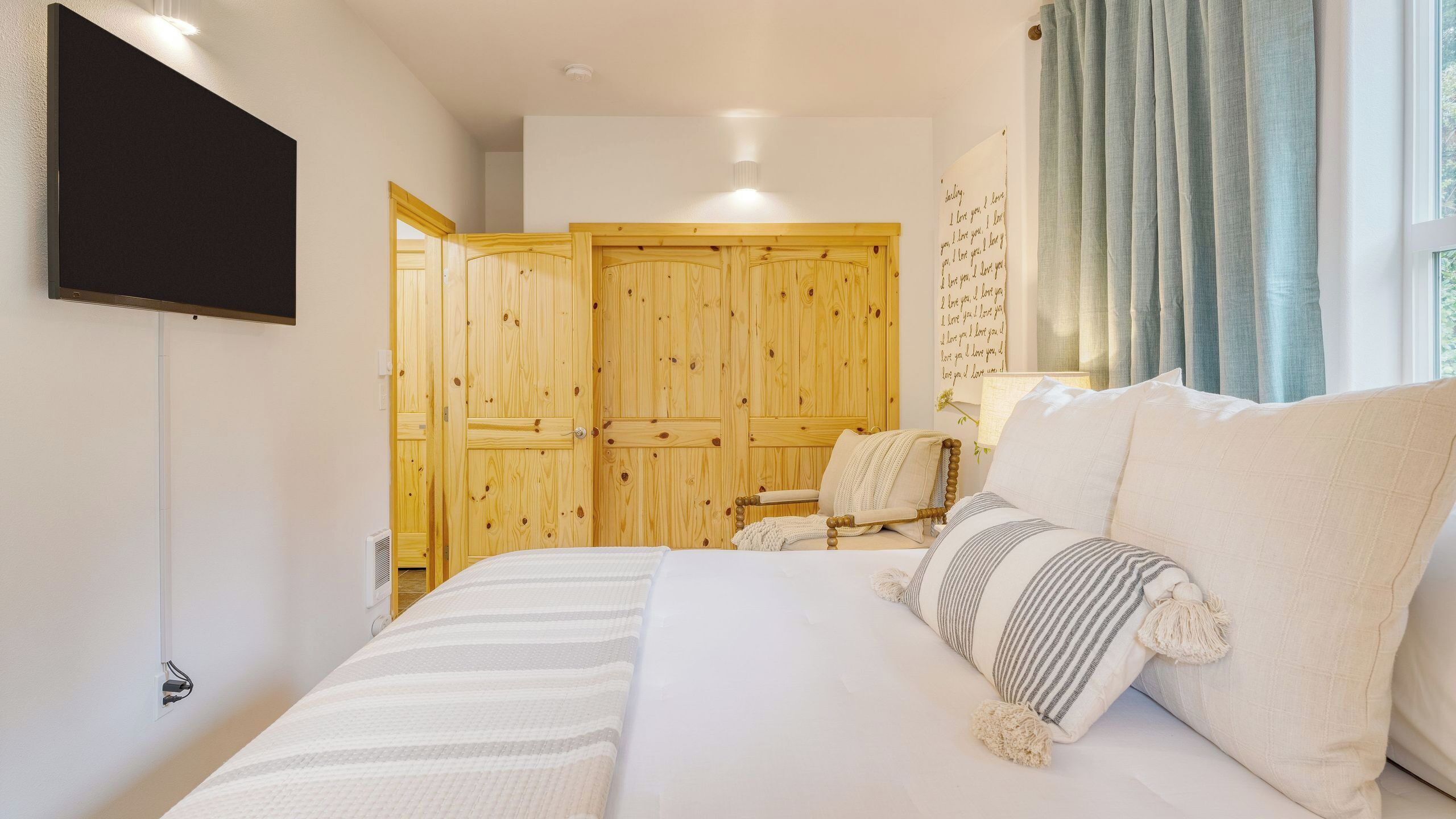 A bedroom with a bed, a wooden wardrobe, a television, and a window with curtains.