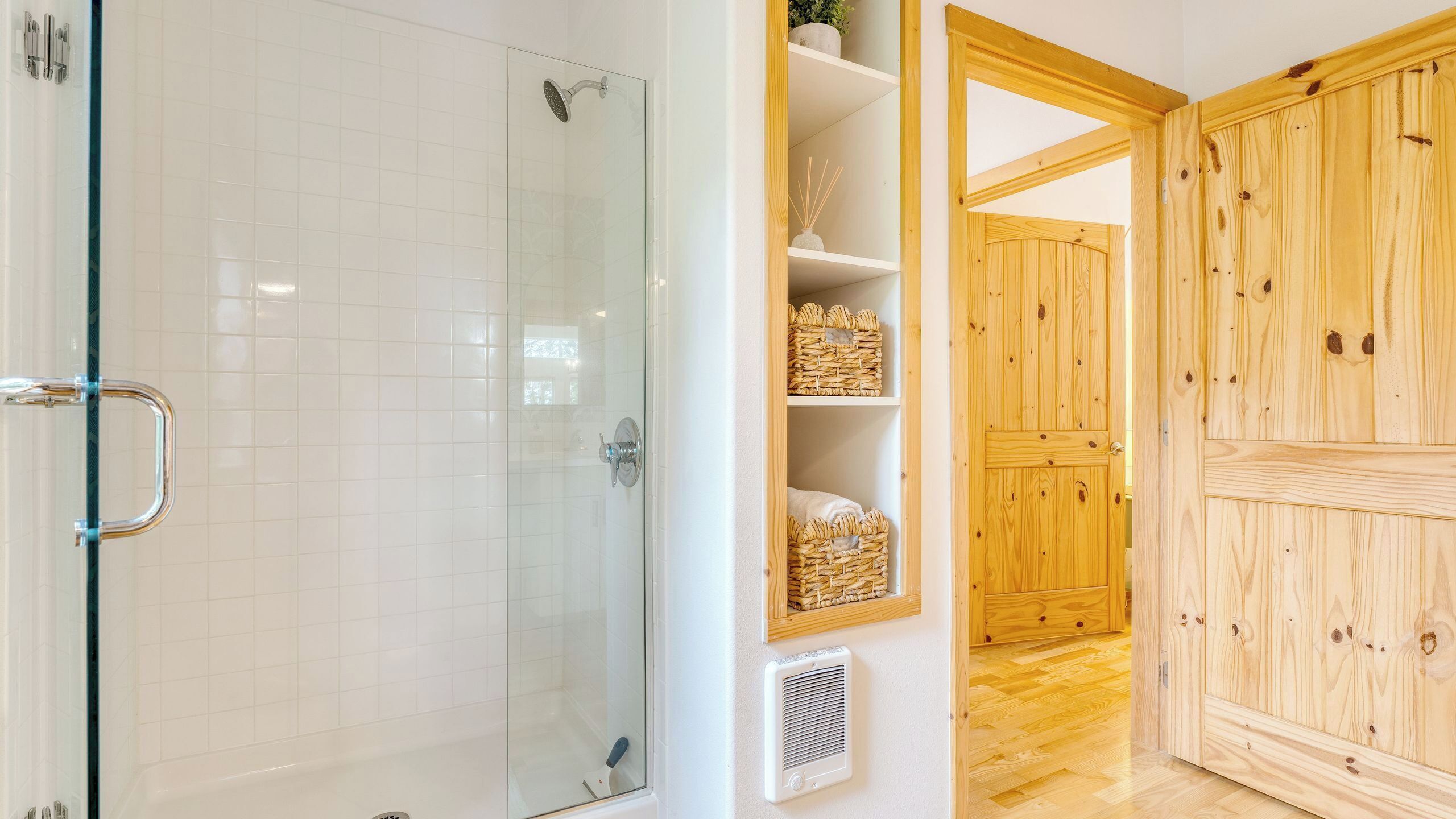 Un baño con cabina de ducha de vidrio, pared de azulejos blancos, puerta de madera y estante con cestas.