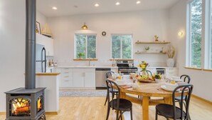 A modern kitchen with a wood-burning stove, white cabinetry, and a dining area with a wooden table and chairs. - Buttercup Bay Hideaway by Avantstay Cozy Escape w/ Fire Pit, Walk to Beach (Manzanita)