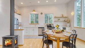 A modern kitchen with a wood-burning stove, white cabinetry, and a dining area with a wooden table and chairs. - Buttercup Bay Hideaway by Avantstay Cozy Escape w/ Fire Pit, Walk to Beach (Manzanita)