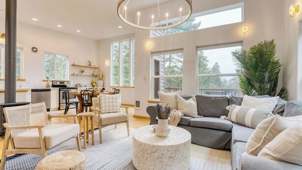 A spacious living room with a fireplace, a dining area, and large windows offering a view of the outdoors. - Buttercup Bay Hideaway by Avantstay Cozy Escape w/ Fire Pit, Walk to Beach (Manzanita)