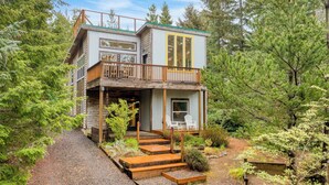 A two-story cabin with a balcony, surrounded by dense forest. - Buttercup Bay Hideaway by Avantstay Cozy Escape w/ Fire Pit, Walk to Beach (Manzanita)