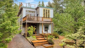 A two-story cabin with a balcony, surrounded by dense forest. - Buttercup Bay Hideaway by Avantstay Cozy Escape w/ Fire Pit, Walk to Beach (Manzanita)