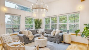 A spacious living room with a grey sofa, a round coffee table, and a large window overlooking a forest. - Buttercup Bay Hideaway by Avantstay Cozy Escape w/ Fire Pit, Walk to Beach (Manzanita)
