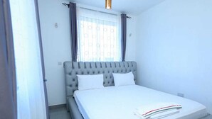 A bedroom with a tufted headboard, a bed with white bedding, a window with curtains, and a light fixture above the bed.