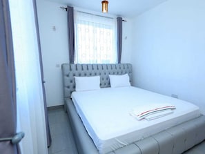 A bedroom with a tufted headboard, a bed with white bedding, a window with curtains, and a light fixture above the bed. - 5 Minutes to the Beach | Stylish 1-Bedroom Apartment in Mombasa (Mombasa)