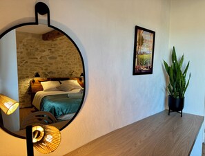 Une chambre avec un mur en pierre, un lit avec une literie blanche, une table de chevet avec une lampe, un tableau encadré au mur et une plante en pot sur une table en bois.