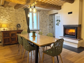 Un espace repas avec une table en bois, des chaises vertes, un mur en pierre, une cheminée et des décorations suspendues.