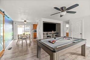 A pool table in a room with a ceiling fan, a dining area with chairs, and a kitchen area with cabinets.