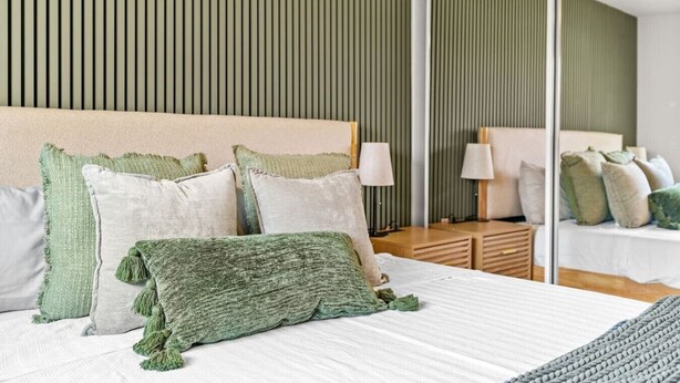 Ein modernes Hotelzimmer mit einem großen Bett, grünen und weißen Kissen, einem Nachttisch mit Lampe und einem Spiegel, der den Raum reflektiert.