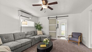 Ein Wohnzimmer mit einem Ecksofa, einem runden Couchtisch, einem Deckenventilator, einer Klimaanlage, einer Schiebetür und einem Sessel.
