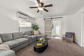 A living room with a sectional sofa, a round coffee table, a ceiling fan, an air conditioning unit, a sliding door, and a chair.