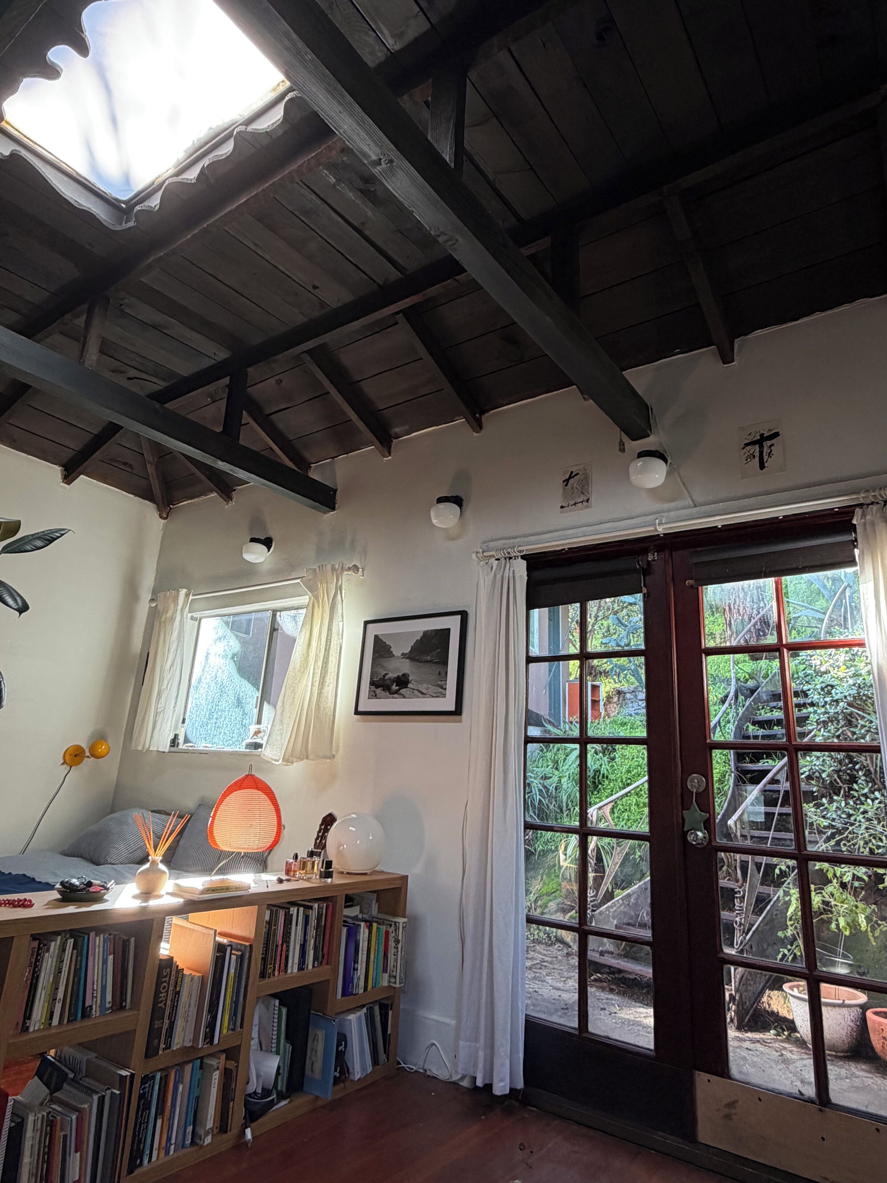 A room with a skylight, wooden ceiling beams, a bookshelf, a desk with a lamp, a framed picture, and a view through a glass door.