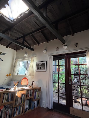 Ein Zimmer mit Oberlicht, Holzbalkendecke, Bücherregal, Schreibtisch mit Lampe, gerahmtes Bild und Blick durch eine Glastür.