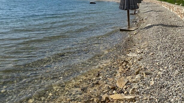 Une plage de galets avec une eau claire, quelques parasols et des bâtiments en arrière-plan.