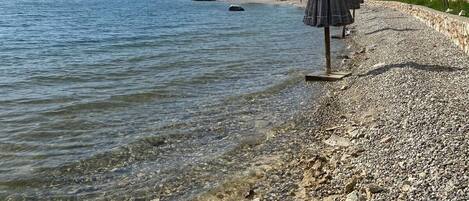 A pebble beach with clear water, a few umbrellas, and buildings in the background.