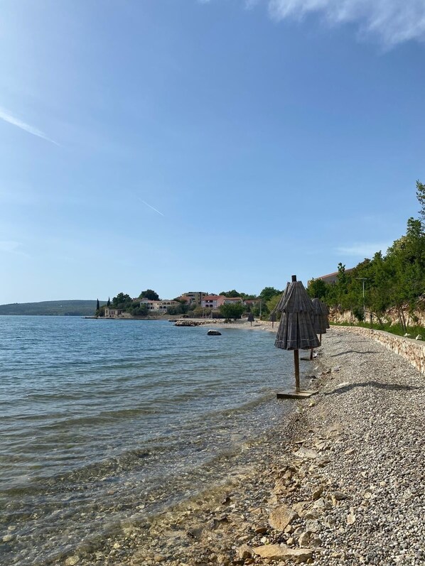 Ein Kiesstrand mit klarem Wasser, einigen Sonnenschirmen und Gebäuden im Hintergrund.