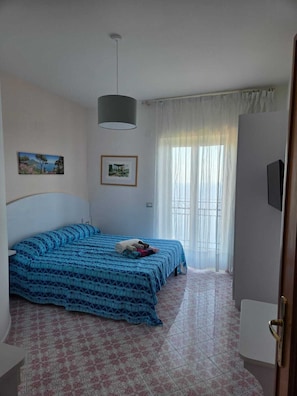 Ein Schlafzimmer mit einem Bett, einer Deckenleuchte, einem Fenster mit Vorhängen und einem an der Wand befestigten Fernseher.