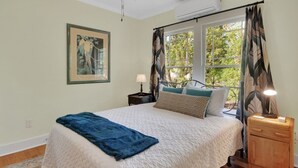 Un dormitorio con cama, mesita de noche, lámpara, ventana con cortinas y un cuadro en la pared.