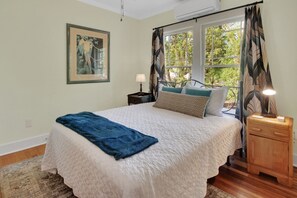 Ein Schlafzimmer mit einem Bett, einem Nachttisch, einer Lampe, einem Fenster mit Vorhängen und einem gerahmten Bild an der Wand.