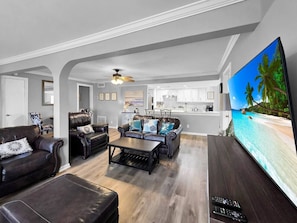 Ein geräumiges Wohnzimmer mit Ledersofas, einem Couchtisch und einem großen Flachbildfernseher, der eine Strandansicht zeigt.