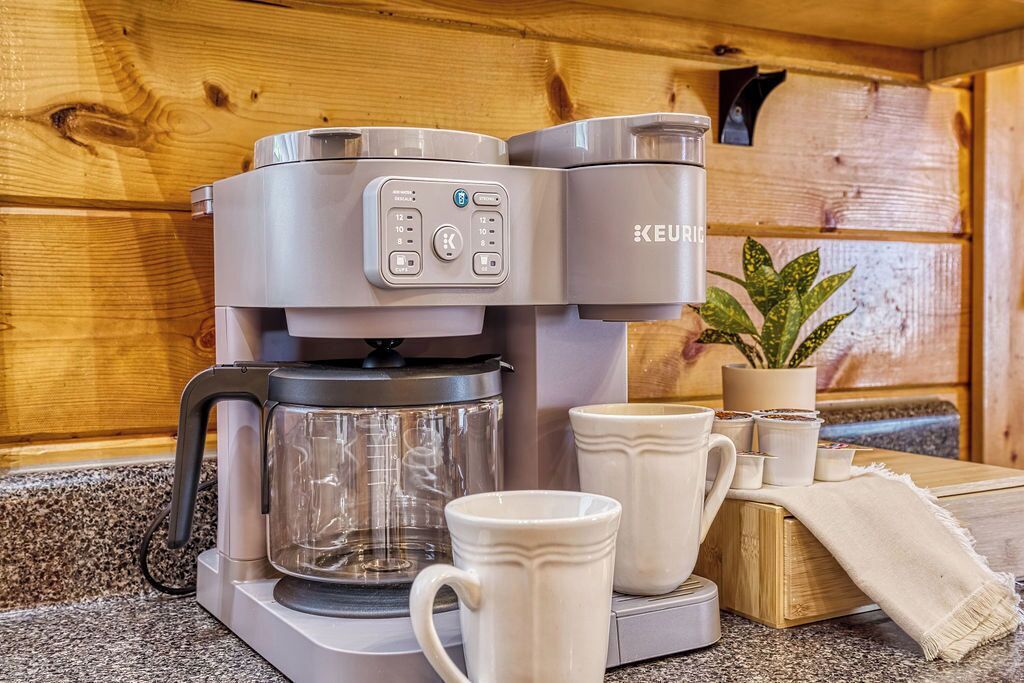 Een Keurig koffiezetapparaat met een glazen kan, twee witte mokken en een kleine plant op een houten aanrecht.