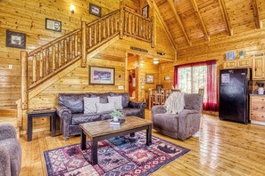 Ein Interieur einer Blockhütte mit einer Holztreppe, einem Wohnzimmer mit einer Couch und Sesseln, einem Essbereich und einer Küche mit einem Kühlschrank.