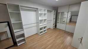 Huoneessa on puulattia, valkoinen vaatekaappi, jossa on useita hyllyjä, sekä peilikaappi.