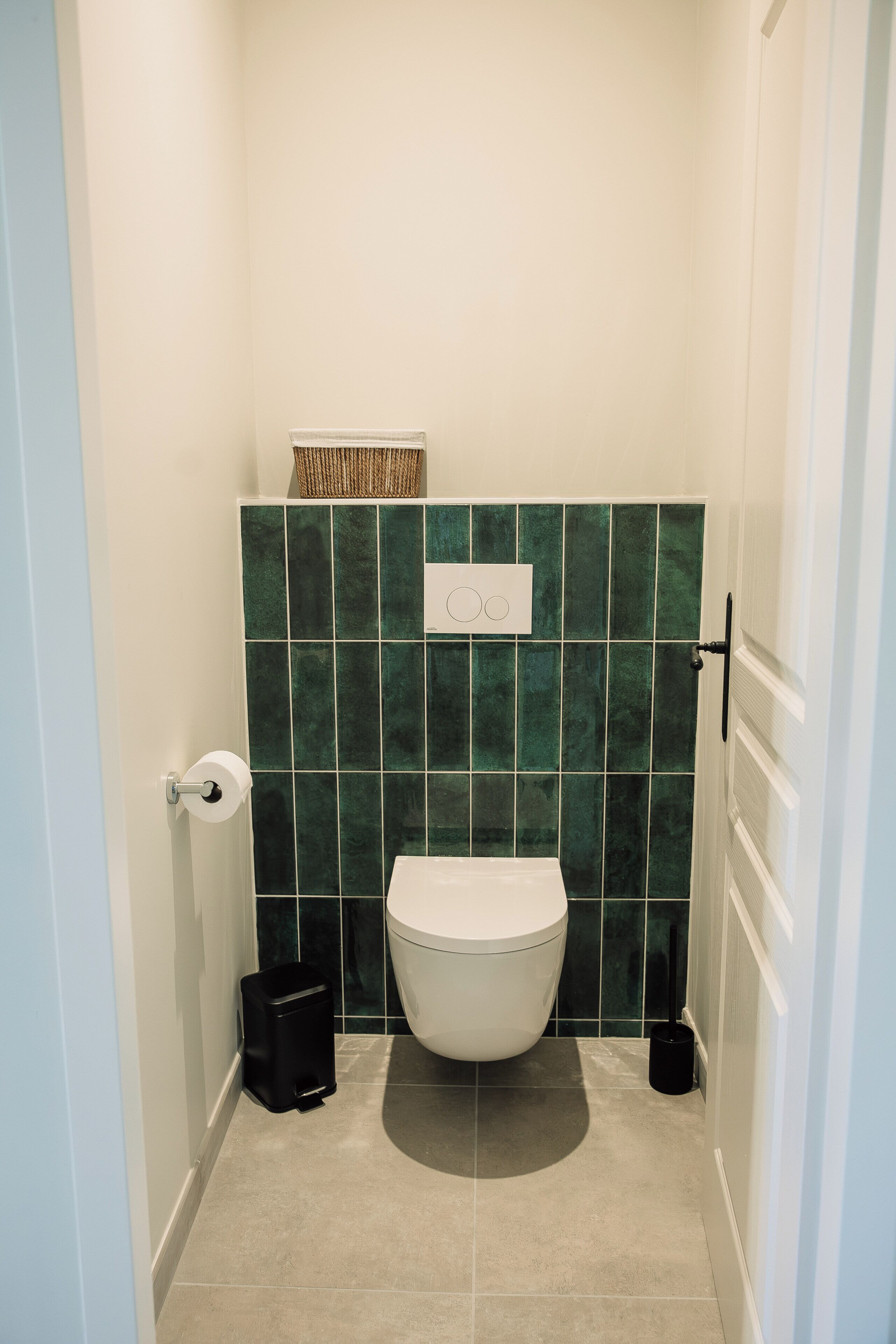 A small, narrow bathroom with a toilet, a trash bin, and a wall-mounted toilet paper holder.
