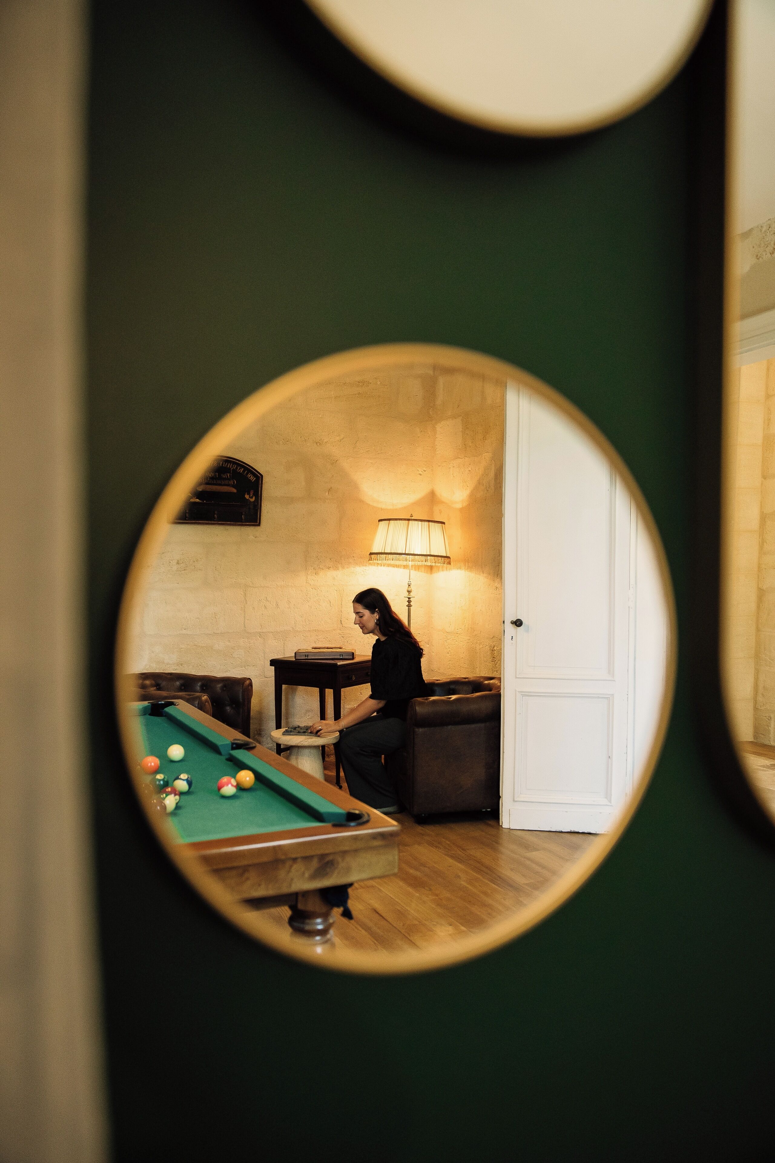 Une table de billard avec des billes, une personne assise à la table, une lampe et une porte dans une pièce aux murs verts.