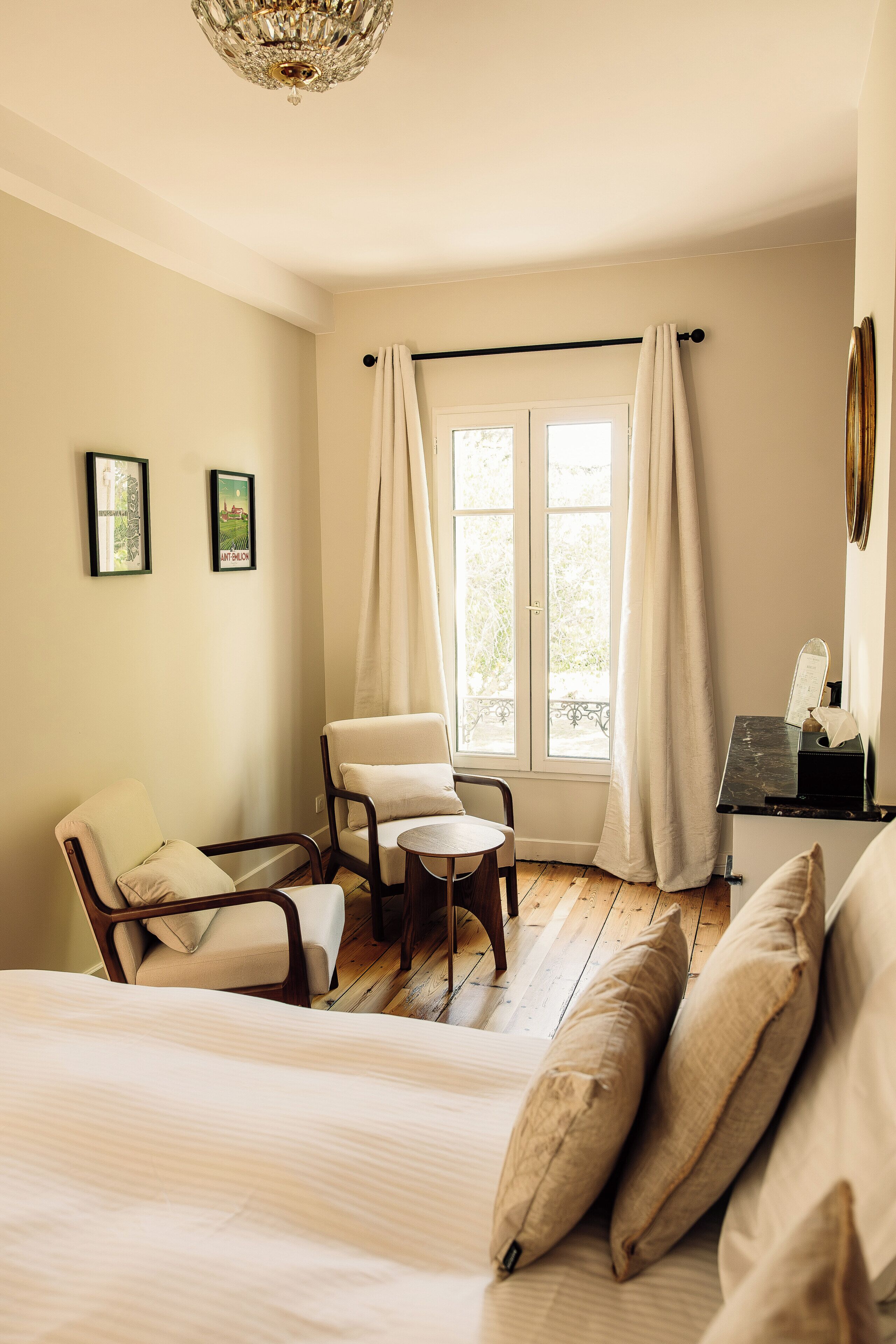 Een kamer met een bed, twee stoelen, een tafeltje, een raam met gordijnen en twee ingelijste foto's aan de muur.