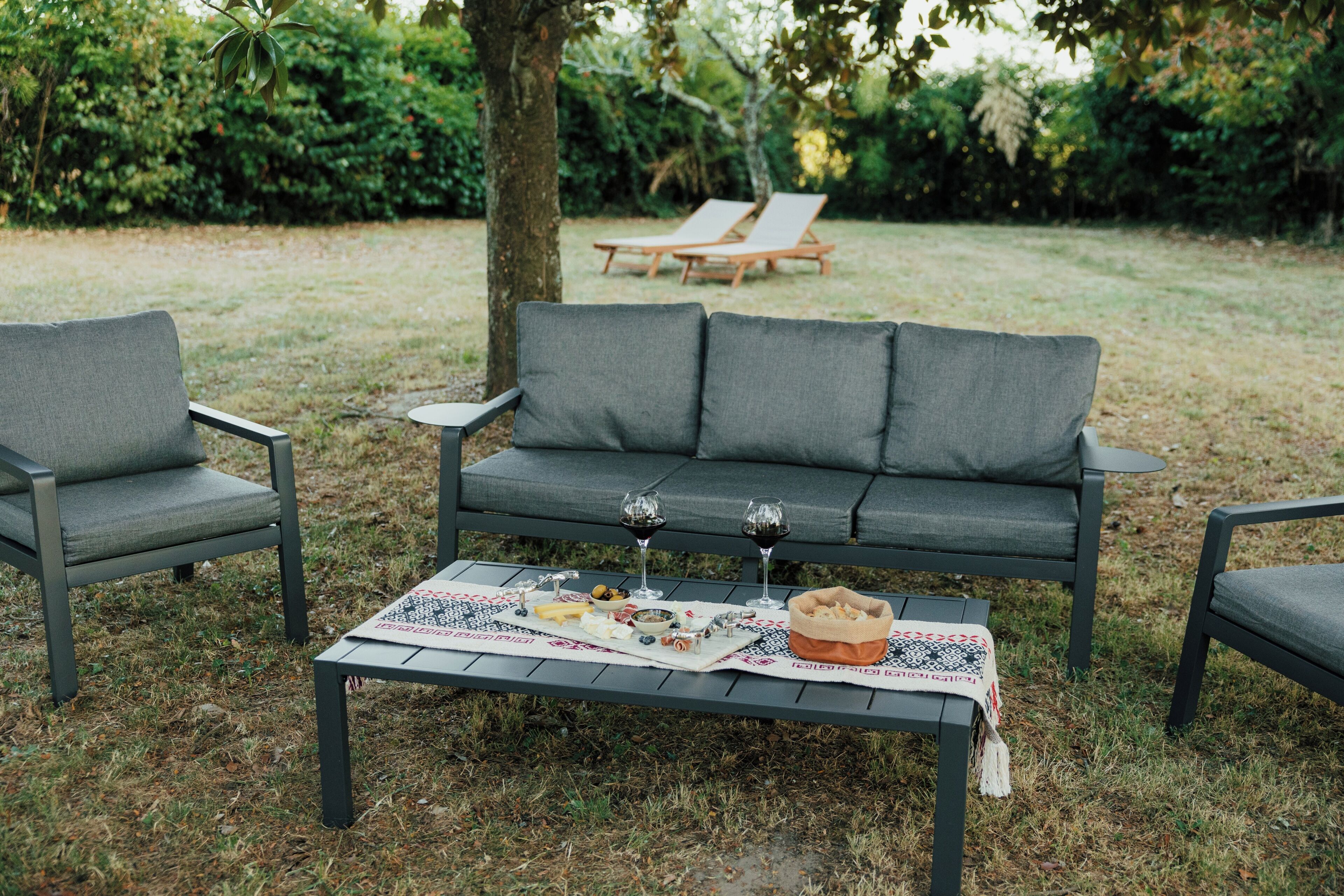 Um jardim com um sofá cinzento, duas poltronas e uma mesa com um jogo de jantar, copos de vinho e comida.