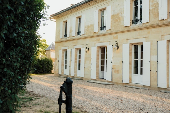 A building with multiple windows and doors, a gravel driveway, and a hedge in the foreground.