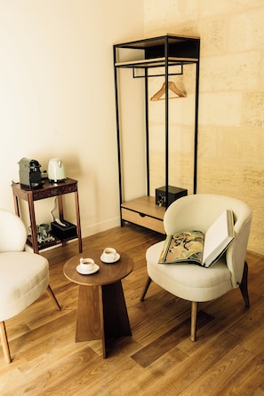 A room with a round wooden coffee table, two armchairs, a small side table with a kettle and cups, and a hanging light fixture.