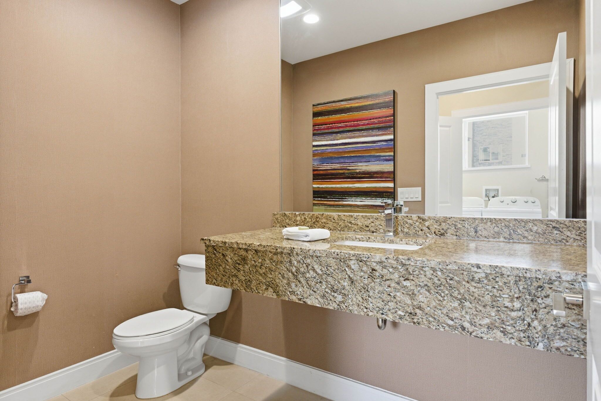 Une salle de bain comprenant des toilettes, un lavabo avec un plan de travail en granit, un miroir et un tableau abstrait aux couleurs vives accroché au mur.