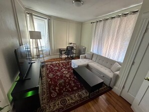 Ein Wohnzimmer mit einer Couch, einem Couchtisch, einem Fernseher, einer Lampe, einem Sessel und einem gemusterten Teppich.