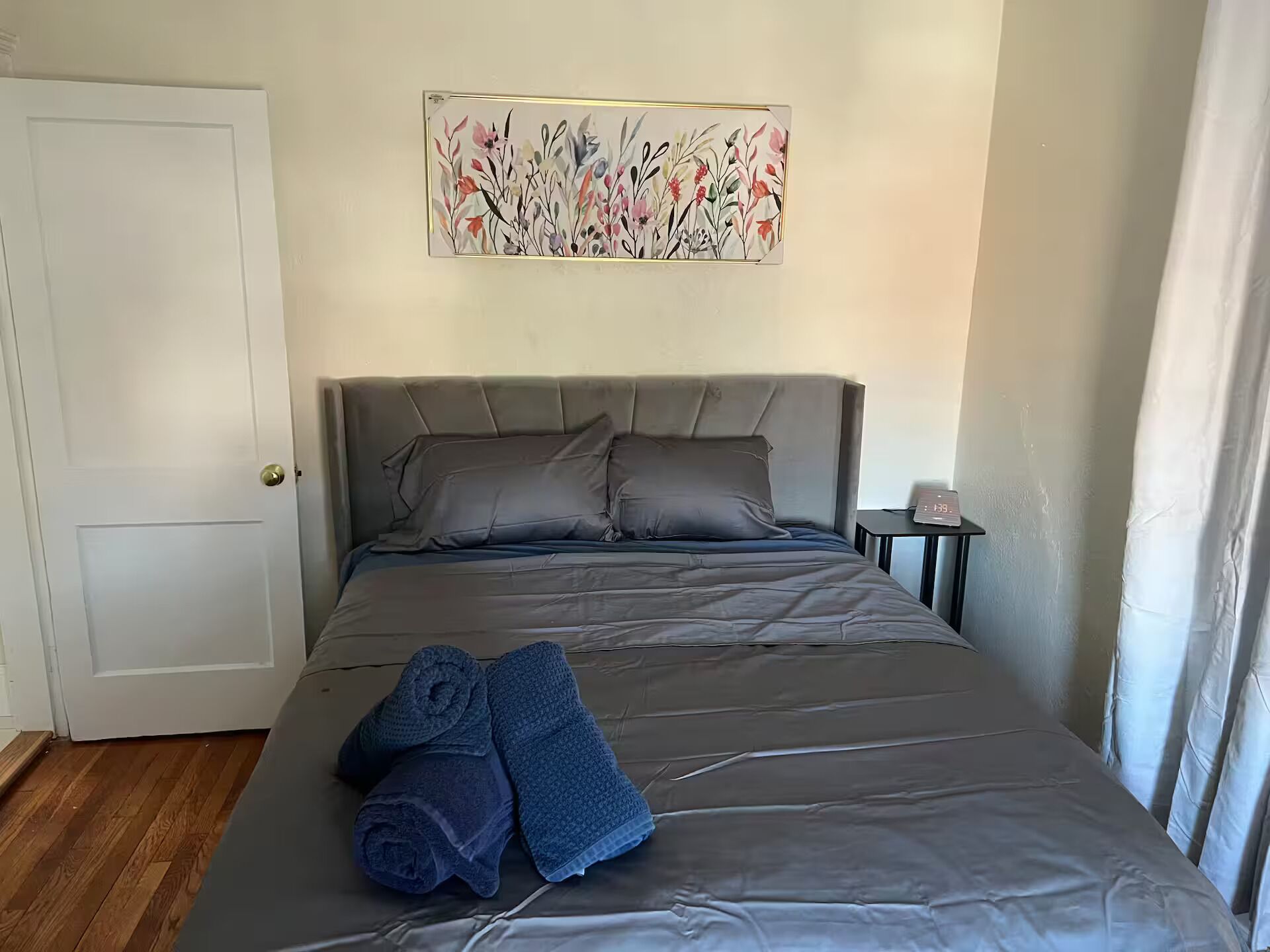 A bedroom with a bed, pillows, a nightstand, a clock, and a painting on the wall.