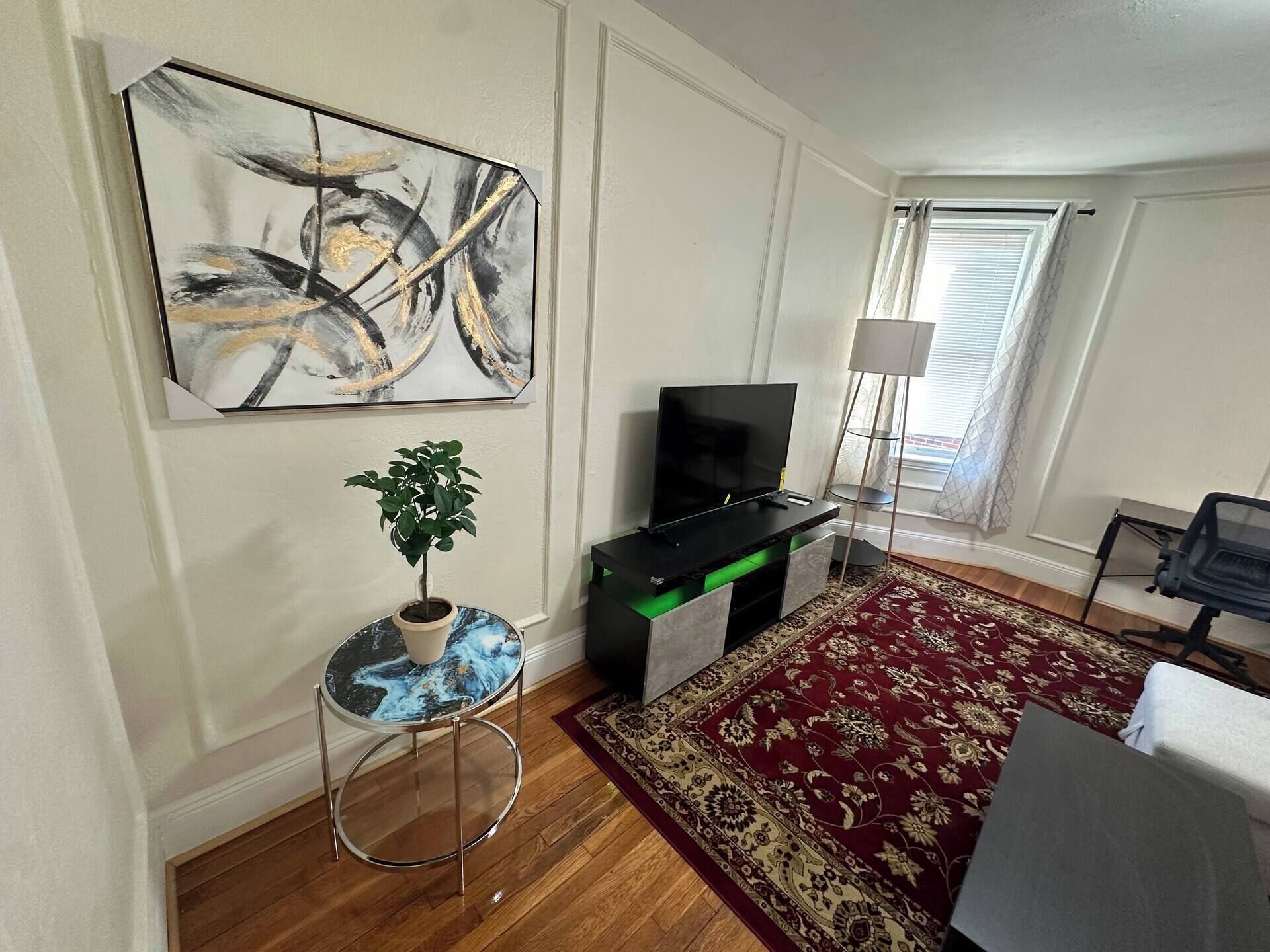 A living room with a flat-screen TV, a potted plant, a round table, a red patterned rug, and a painting on the wall.