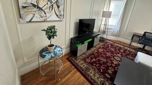 Ein Wohnzimmer mit Flachbildfernseher, Blumentopf, rundem Tisch, farbenfrohem Teppich und einem Bild an der Wand.