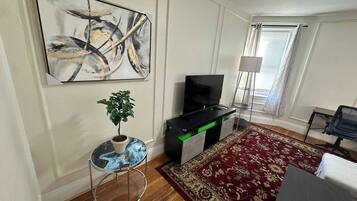 A living room with a flat-screen TV, a potted plant, a round table, a red patterned rug, and a painting on the wall.