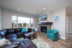 Un salon doté d’un canapé d’angle en cuir, d’un fauteuil turquoise, d’une cheminée, de grandes fenêtres et d’une vue sur les montagnes.